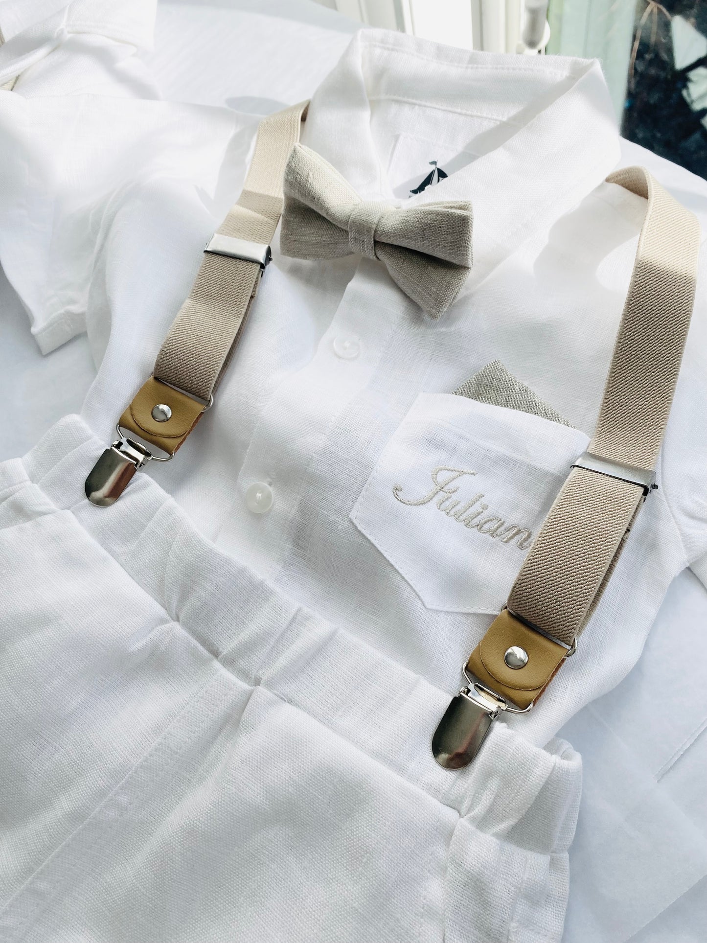 White linen shirt, pants, and beige bowtie, beige suspenders, beige/gold embroidered name Julian on pocket.