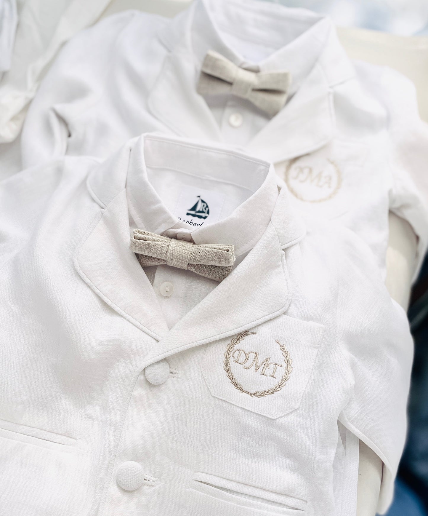 White linen suit with jacket, shirt, pants, and beige linen bowtie, gold stitching on jacket pocket, the initials DMT in monogram form.