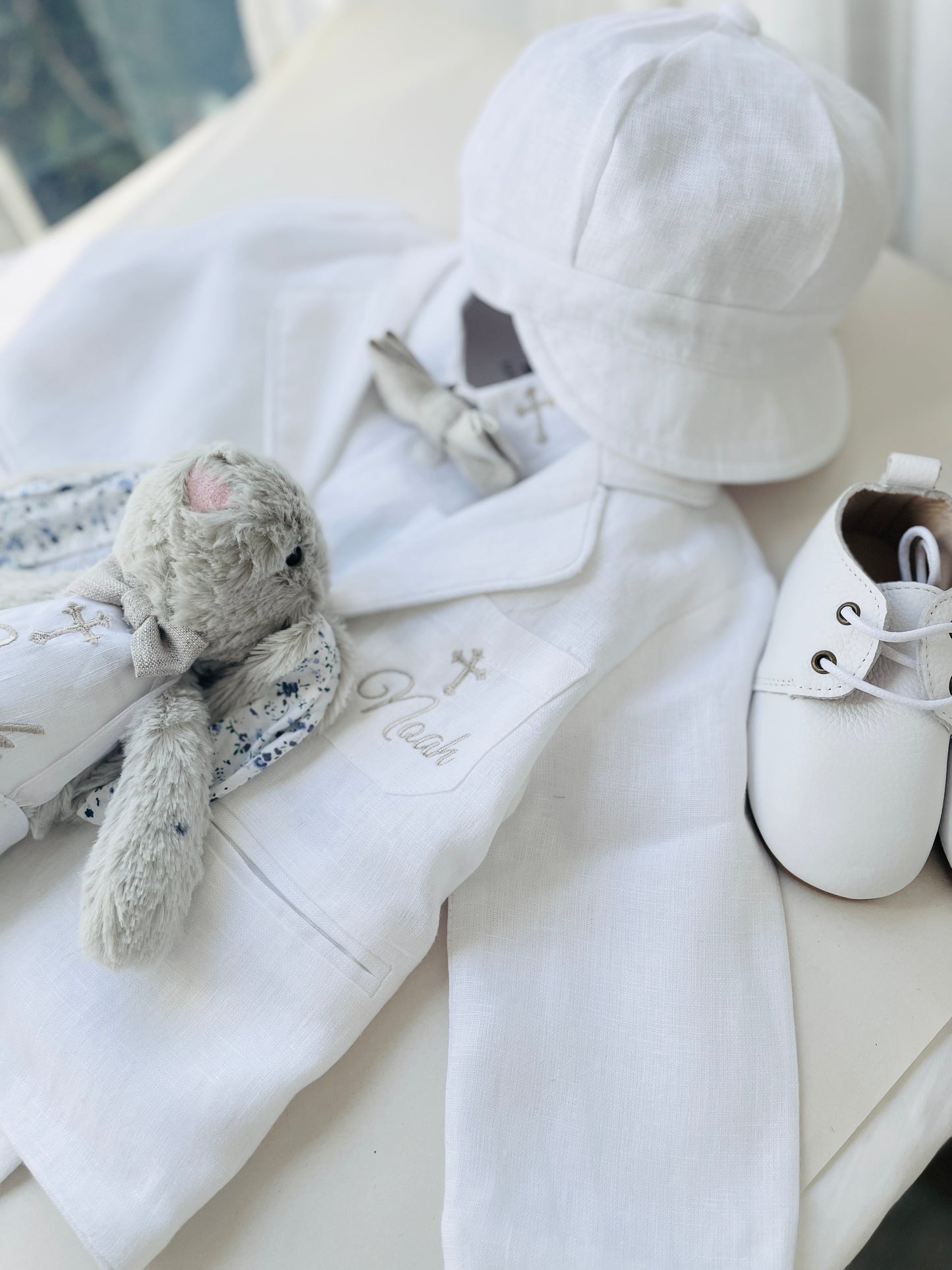 White linen suit with jacket, shirt, pants, and white bowtie, gold stitching on jacket pocket, the name Noah, and a cross above. White leather shoes, white linen cap, grey bunny. Beige bowtie