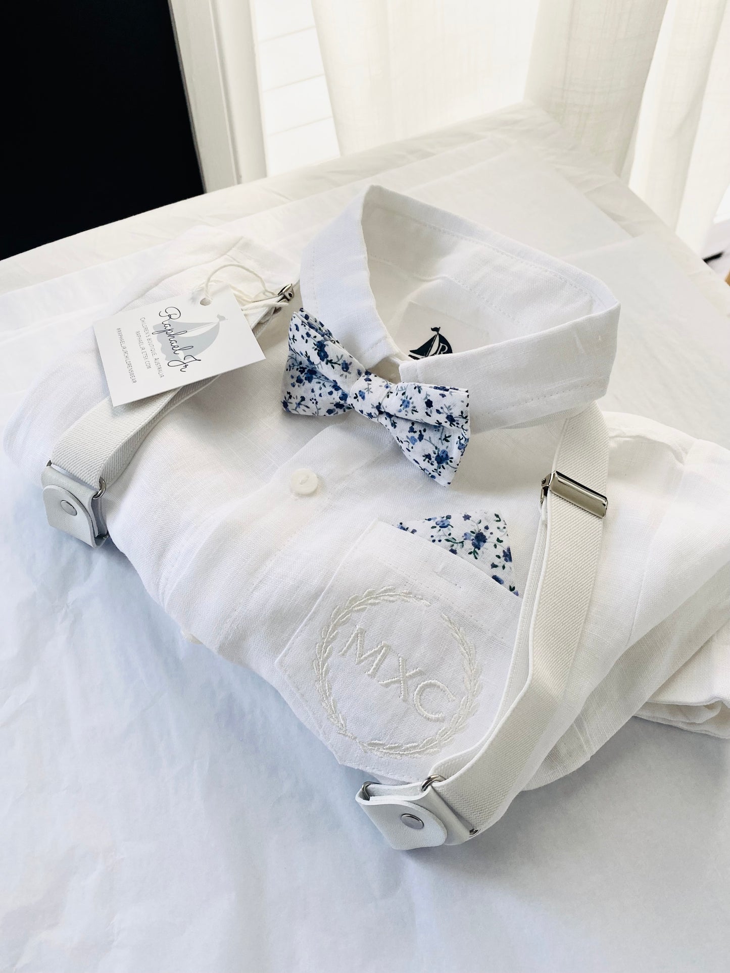 White linen shirt, pants, and white and blue floral bowtie, white suspenders. Stitching of initials MXC with a border, in white thread on shirt pocket.