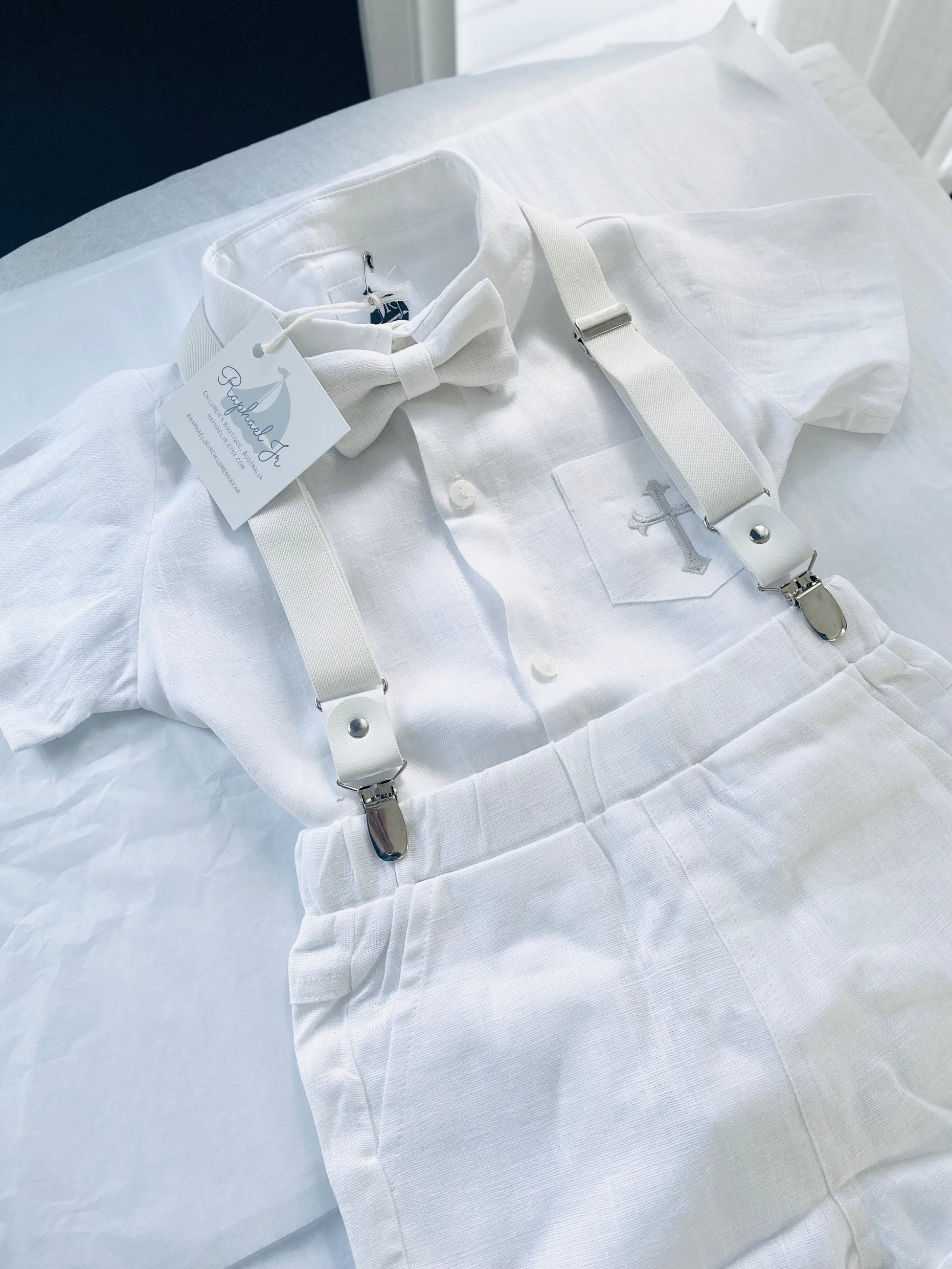 White linen shirt, shorts and white bowtie, light grey cross stitched on pocket, white suspenders.